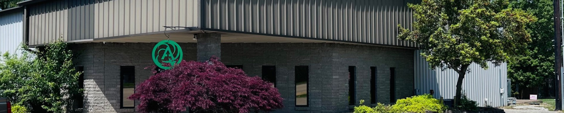 A brick building with vertical metal siding and a green circular logo, surrounded by lush trees and purple bushes.