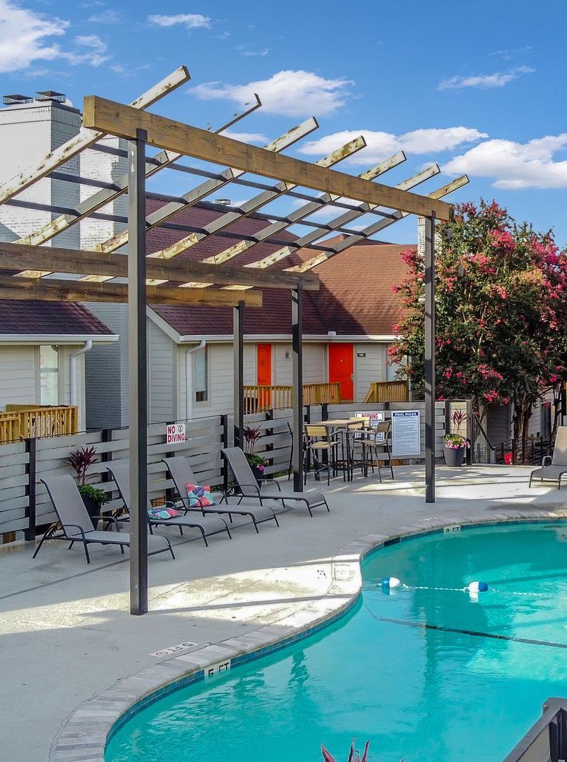 Poolside area with lounge chairs, pergola, and pool with turquoise water. Buildings are visible in the background.