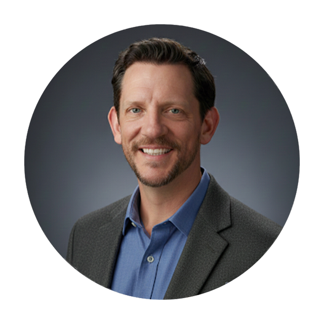 Smiling man in a blue shirt and grey jacket, with a dark background.