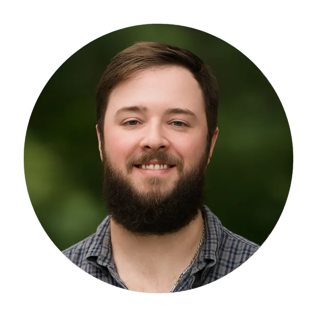 Man with a beard smiling, wearing a plaid shirt, against a blurred green background.