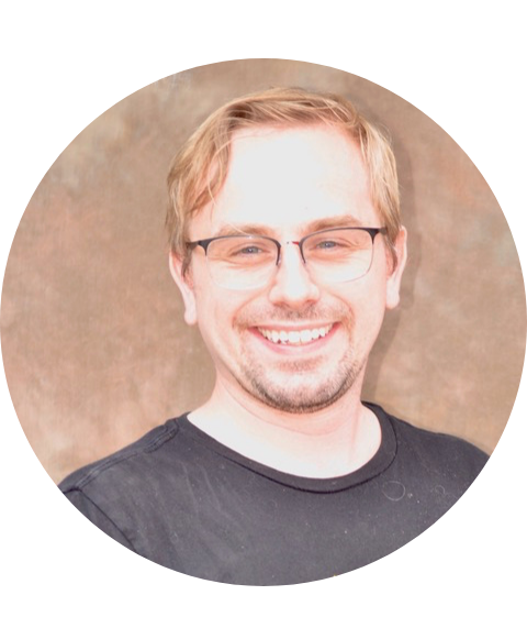 Man with glasses smiling, black shirt, against a blurred brown background.