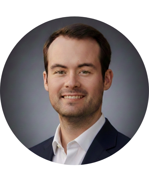 Man with dark hair and a smile wearing a suit jacket over a white shirt, on a gradient background.