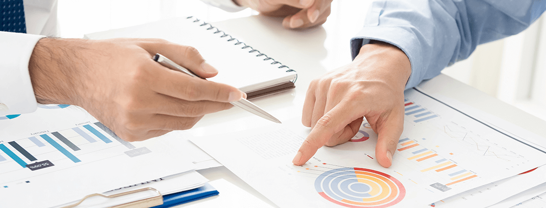 Two people reviewing charts and graphs, one pointing with a finger. They are in a bright office setting.