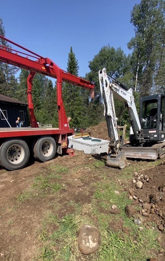 Crane lifting a septic tank near an excavator on a construction site, surrounded by trees and dirt.