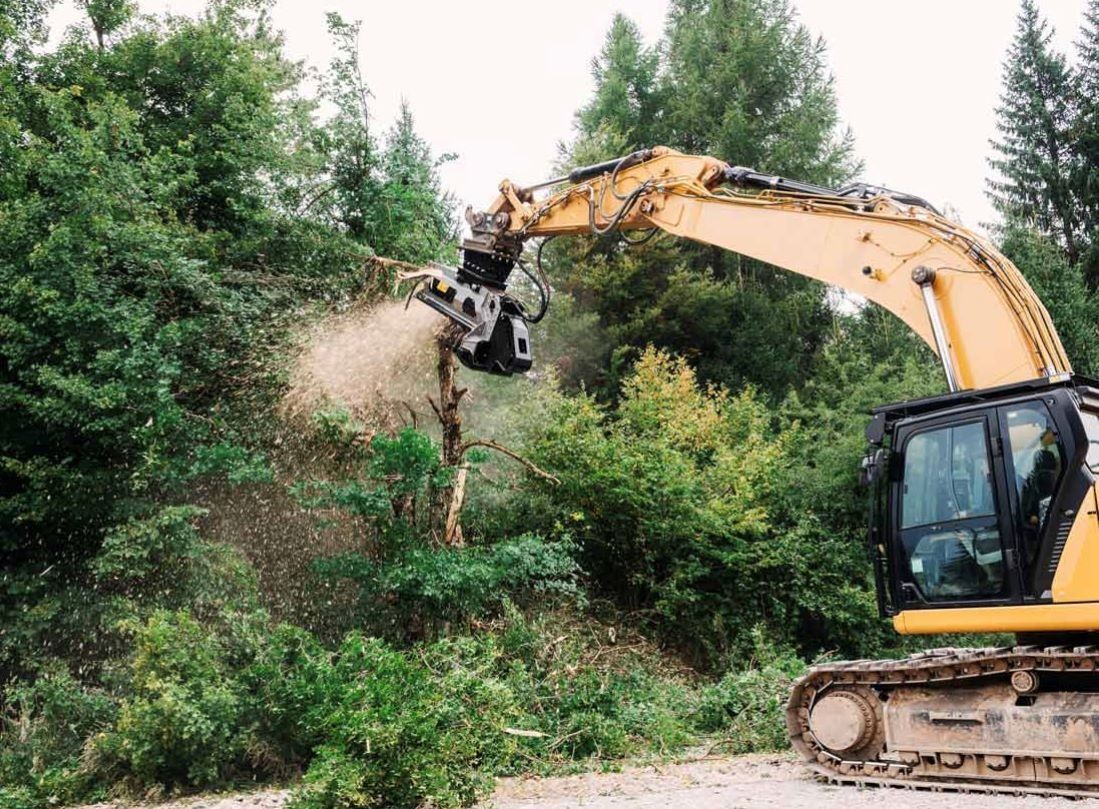 Yellow excavator mulching trees in a forested area, spraying wood chips.