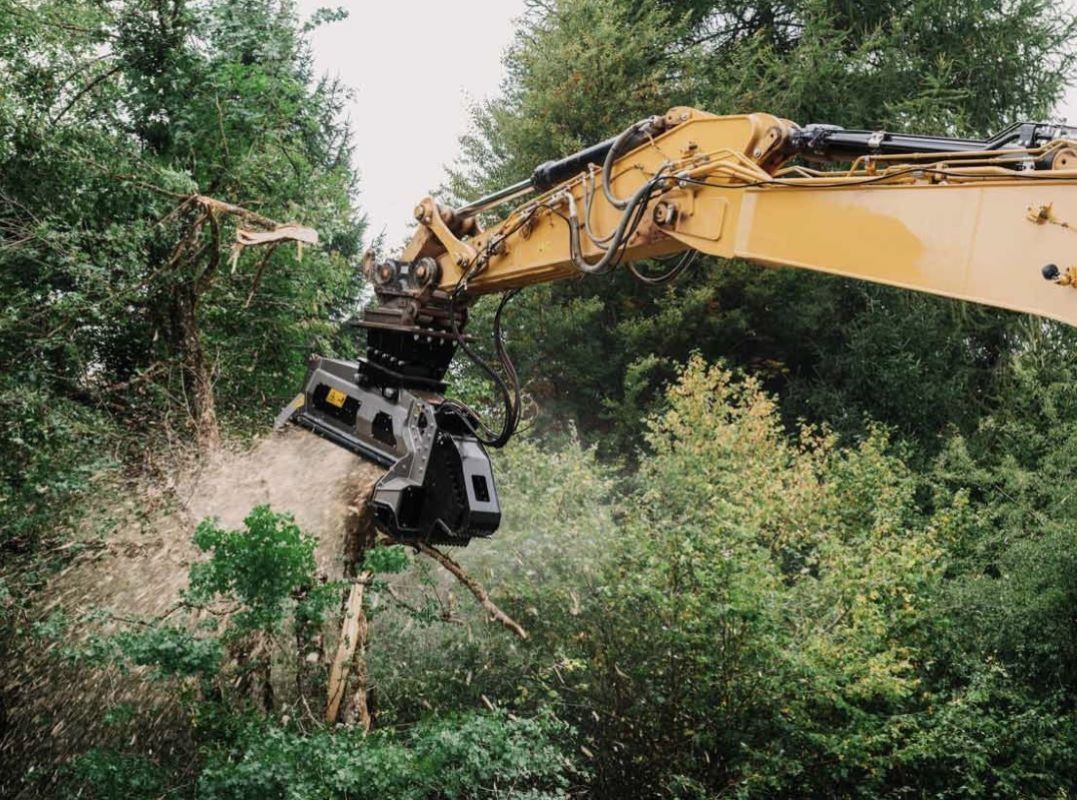 Yellow excavator mulching trees in a forest, wood chips flying.