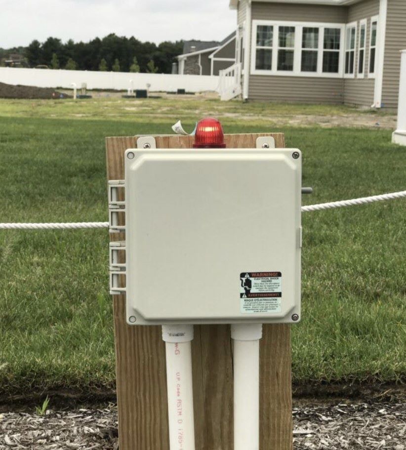 A gray electrical box with a flashing red light mounted on a wooden post in a yard.