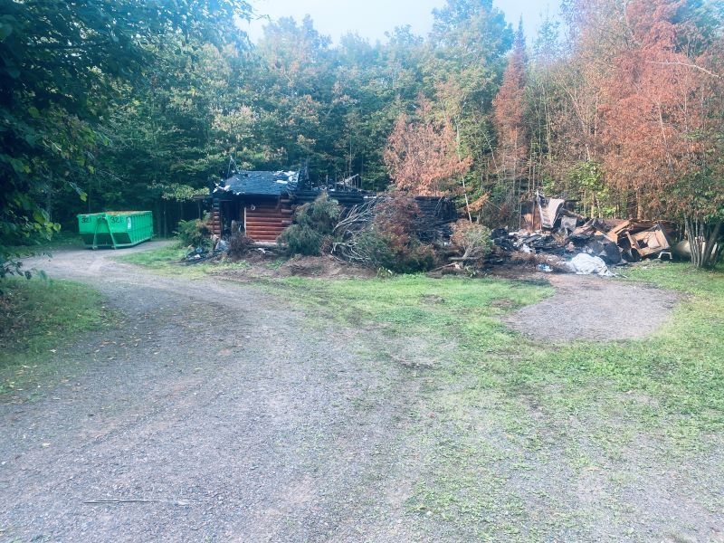 Gravel road leads to a small wooden building in a wooded area with a green dumpster.