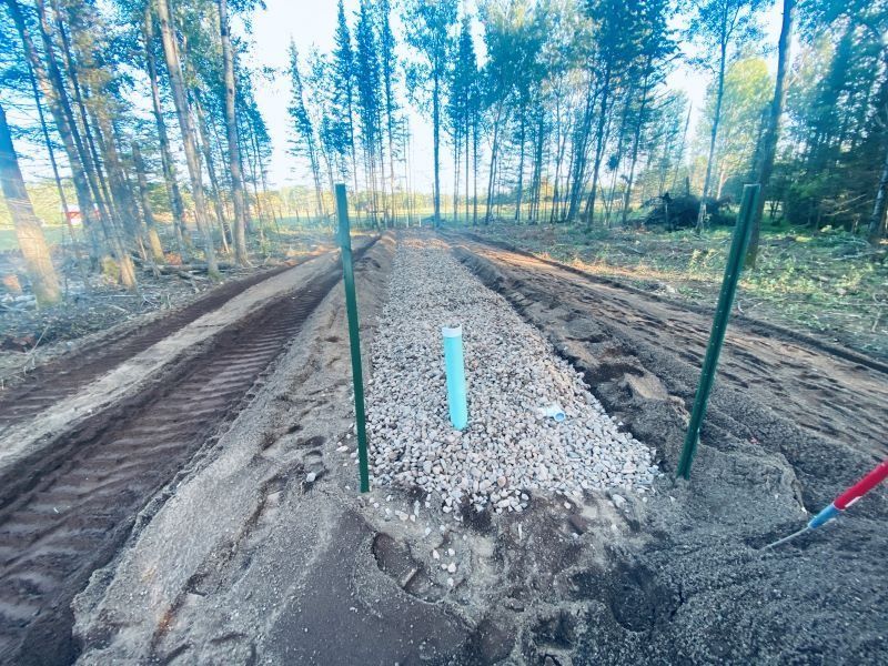 A gravel bed with a blue pipe, between green stakes, in a wooded area, showing construction progress.