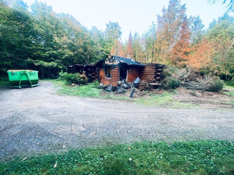 A small log cabin charred by fire, green dumpster, person near wreckage, surrounded by trees.