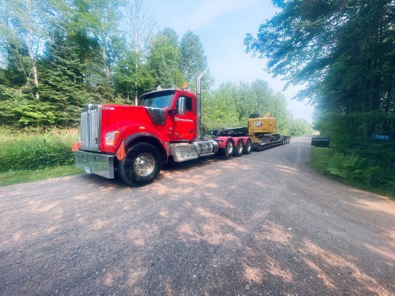 Red semi-truck transporting yellow construction equipment on a gravel road, surrounded by trees and greenery.