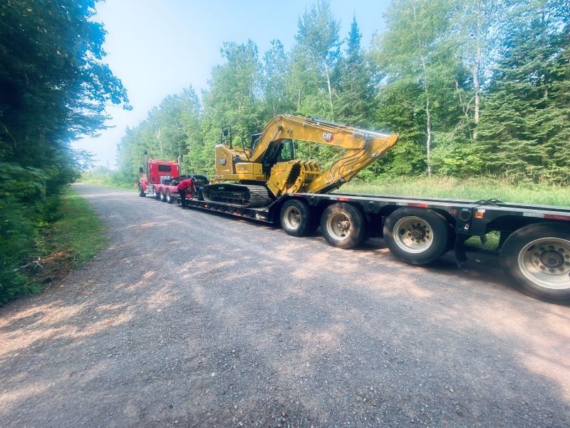 A yellow excavator on a flatbed trailer is pulled by a red semi-truck on a gravel road.