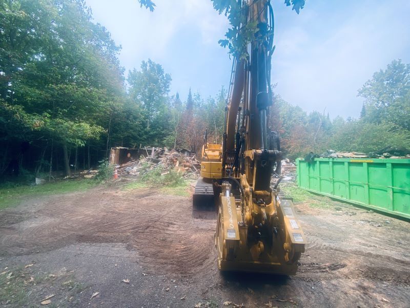 Yellow excavator at work, clearing debris with a green dumpster and trees in the background.