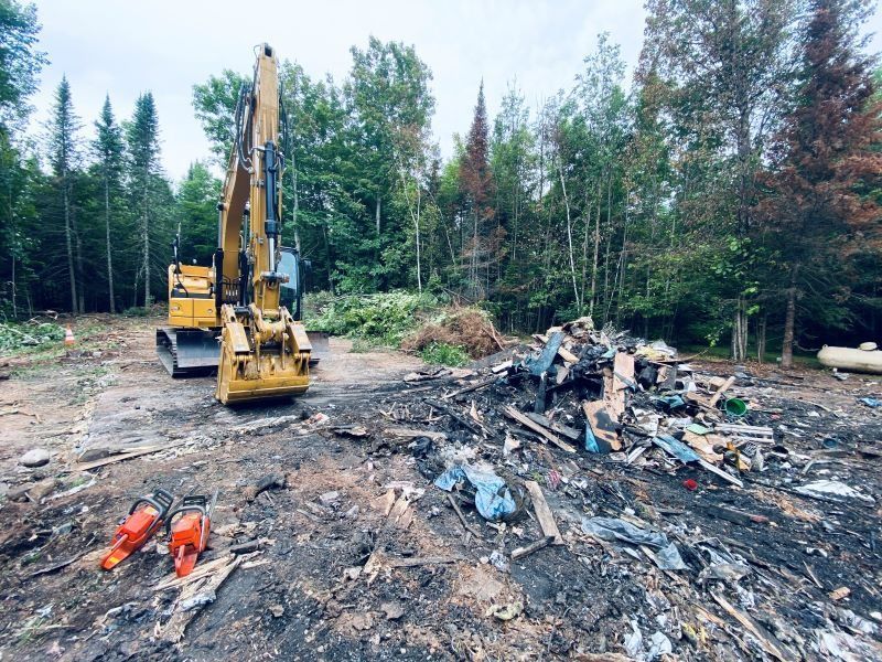 Yellow excavator clearing debris in a wooded area, with a pile of trash and chainsaws in the foreground.