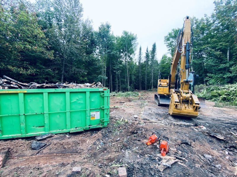 A green dumpster and yellow excavator in a cleared forest area, with a chainsaw on the ground.