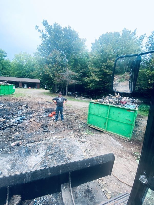 Man standing near a green dumpster. Debris-filled lot, with a building in background. Overcast sky.