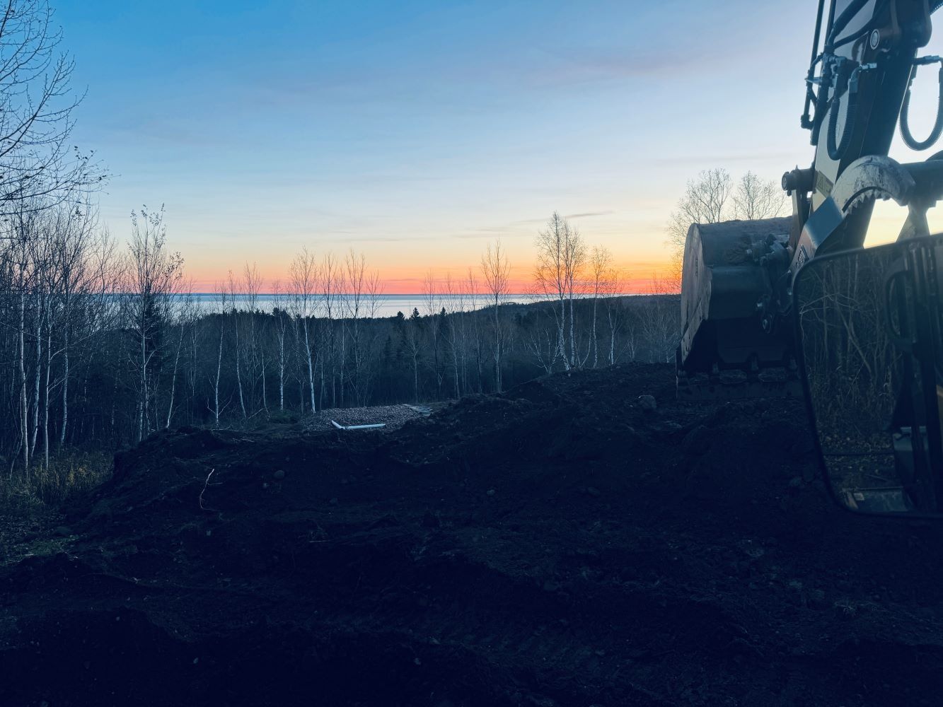 Excavator bucket scoops dark soil, silhouetted against a colorful sunset over a forest and water.