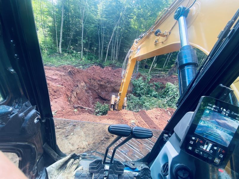 View from excavator cab. Excavator digging into a hillside. Green trees behind the red soil.