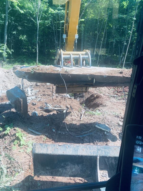 An excavator lifting a large, weathered wooden object in a wooded area.