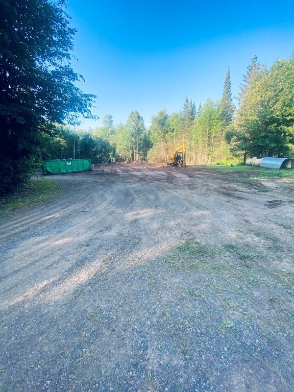 Gravel driveway leading to a clearing in the woods, with a green dumpster and construction equipment.