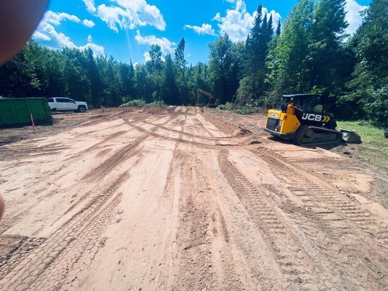 A dirt clearing with tire tracks and a JCB skid steer. Trees in the background, blue sky above.