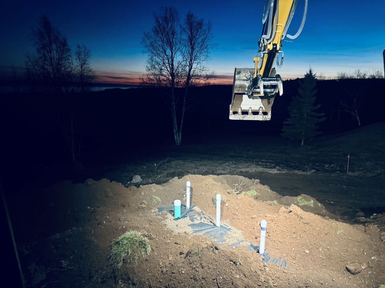 Excavator arm over a construction site at dusk, with pipes and dirt illuminated by the machine's lights.