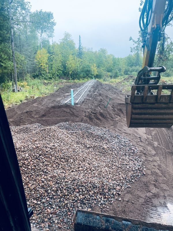 Excavator near a pile of rocks and dirt, with pipes in the ground, likely for a construction project in a wooded area.