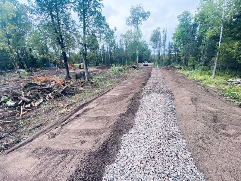 A partially constructed gravel road through a forest. Brown dirt on the sides, a pile of gravel in the center.