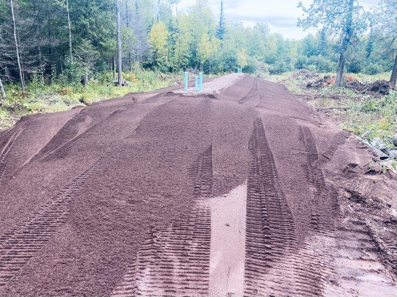 Dirt pile in a wooded area, with tire tracks and blue pipes.