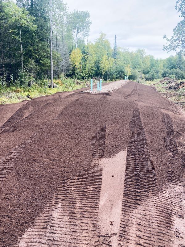 A long mound of dark soil, with tire tracks, in a clearing near a forest.