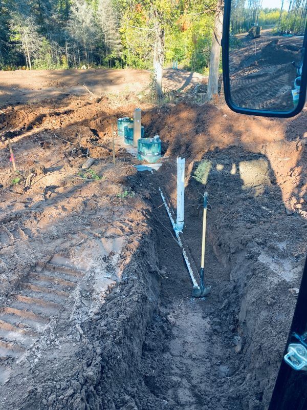 Trench dug in dirt, with pipes and tools visible, from inside of a machine on a construction site.