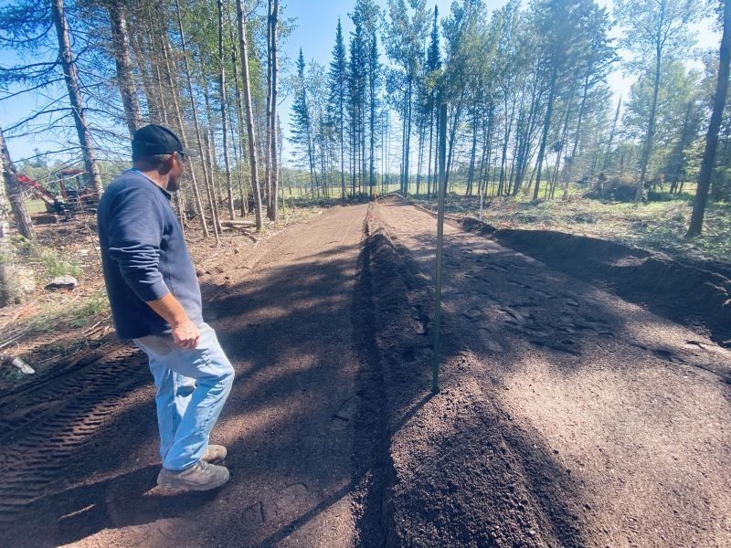 Man in blue shirt and jeans surveys newly graded dirt trail in a forest.