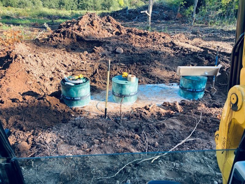 Three green septic tanks in a dirt pit, next to an excavator, ready for installation.