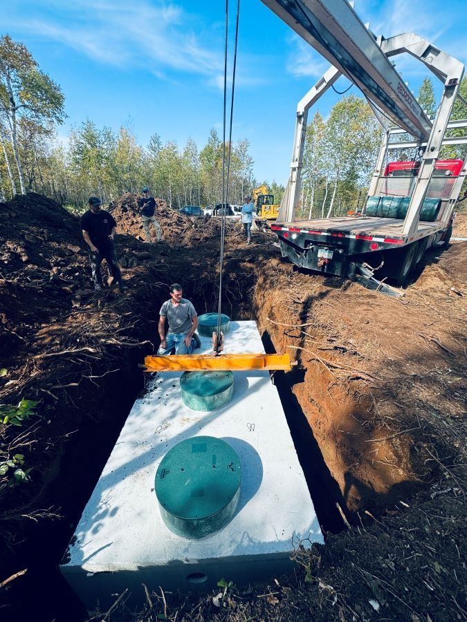 Workers installing a concrete septic tank into a trench on a sunny day. A crane lowers the tank.