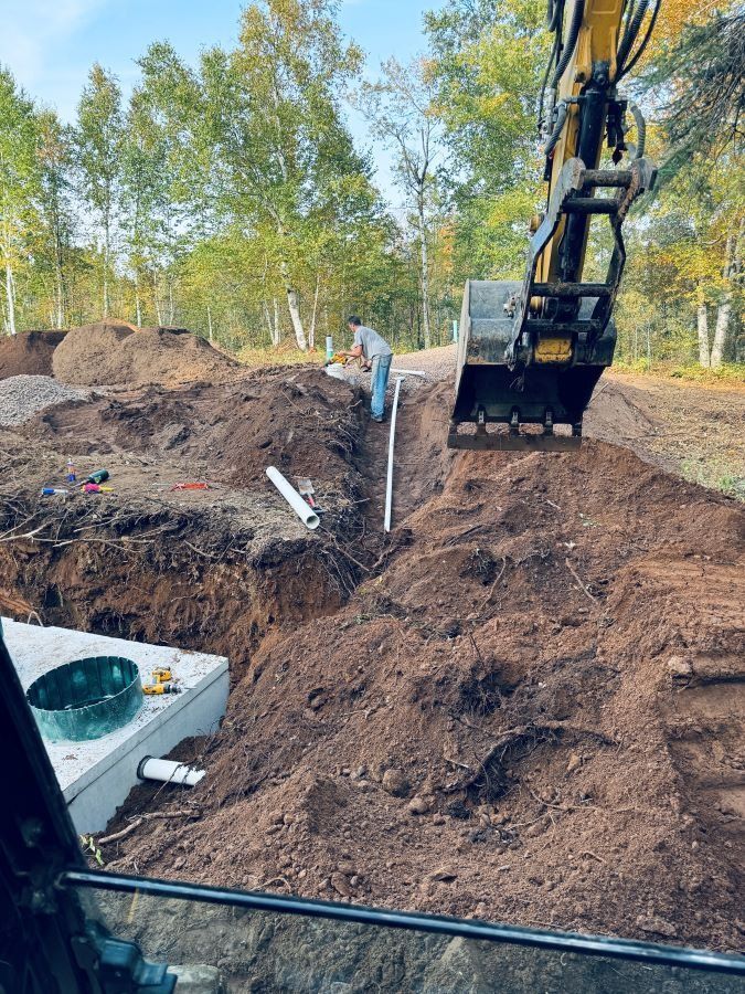 Construction site with a trench, backhoe, and a worker installing pipes in a forest setting.