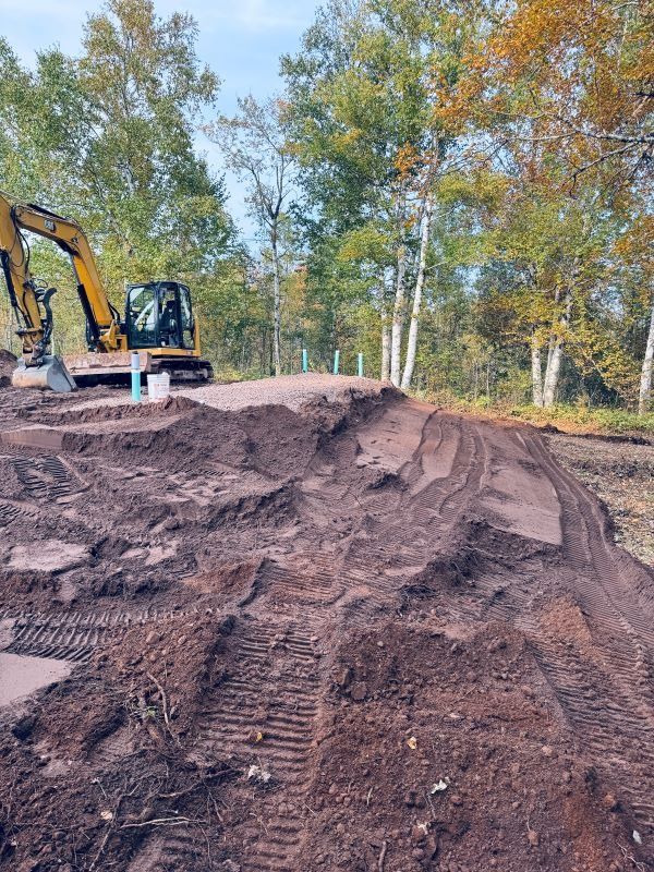 Dirt mounds with an excavator in a wooded area, possibly for construction.