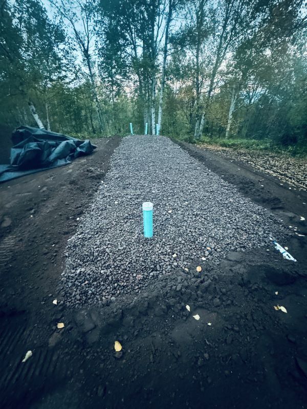 Gravel-covered ground with PVC pipe, bordered by dark soil, trees in the background.