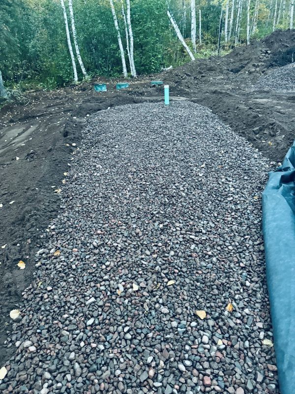A gravel bed with a vertical pipe, surrounded by dirt and trees in the background.