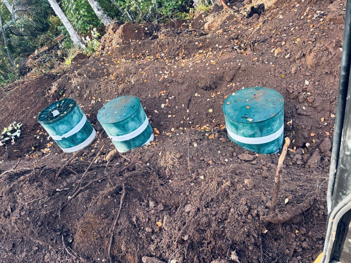 Three cylindrical, teal-colored objects wrapped with white bands, in a dirt clearing, near an excavator.