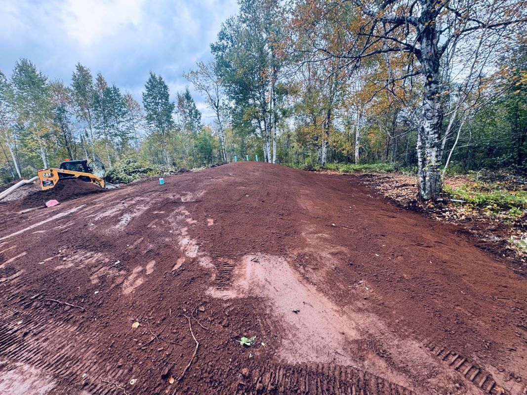 A dirt mound in a wooded area, with an excavator in the background, under an overcast sky.