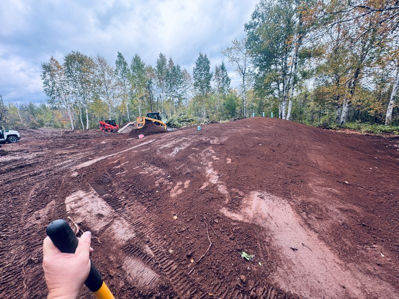 Person holding a shovel, dirt mound in the foreground. Construction site with heavy machinery and trees in the background.