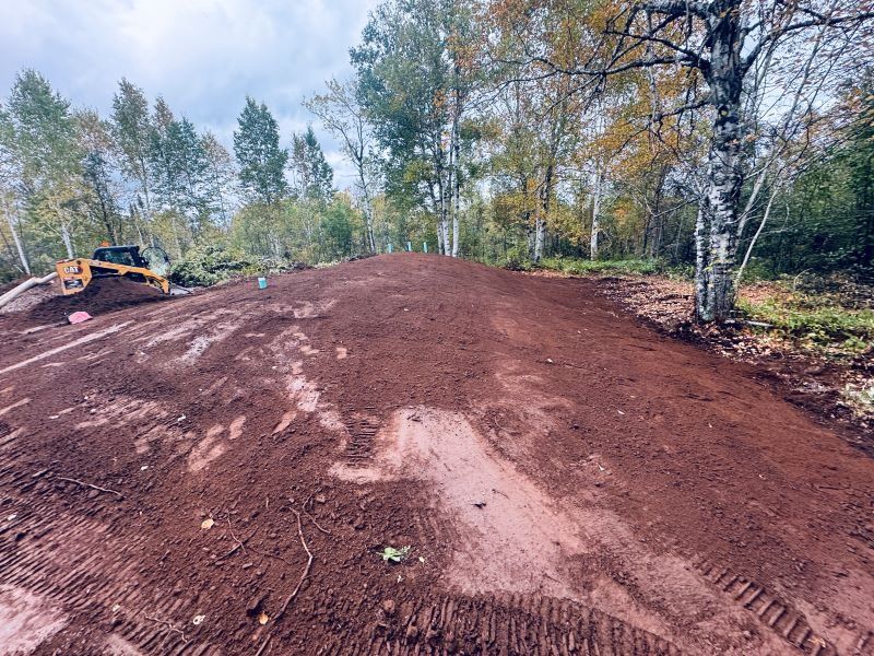 A dirt bike track in progress, with a bulldozer on the left, trees in the background, and red soil.