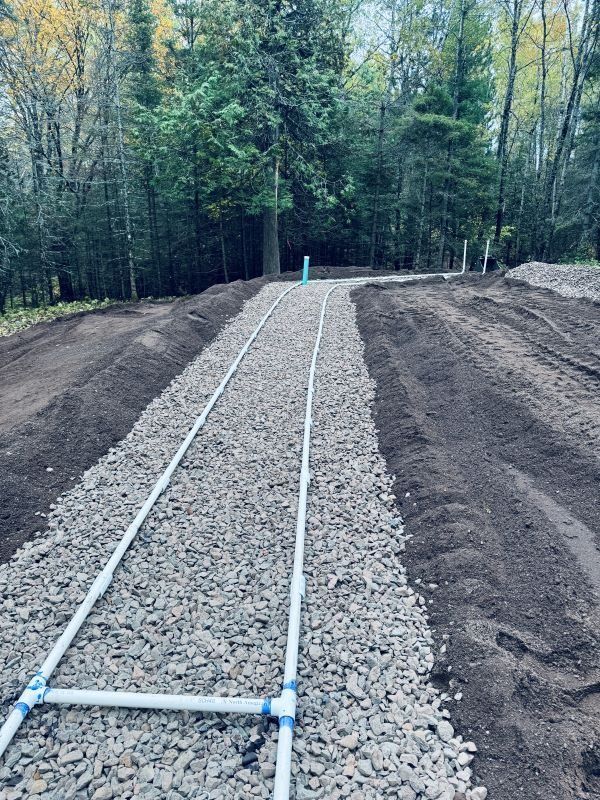 Gravel-filled trench with white pipes, possibly for drainage, surrounded by dirt, in a wooded area.