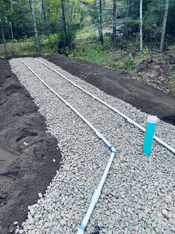 Gravel-filled trench with white pipes and a blue vent pipe; part of a septic system in a wooded area.