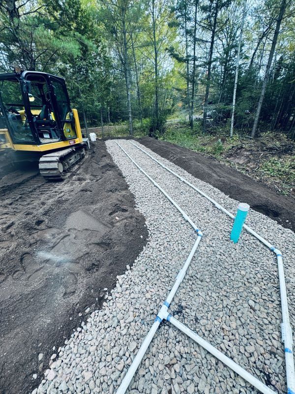 Construction site with gravel bed, white pipes, and yellow excavator in wooded area.
