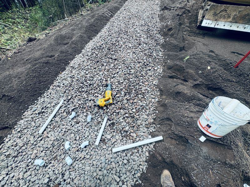 Gravel-filled trench with white PVC pipes, power drill, and bucket on a hillside.