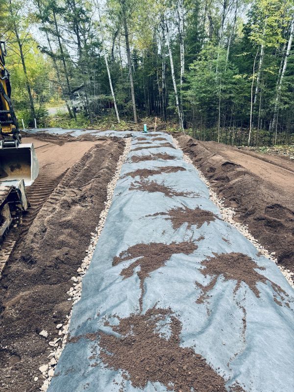 Black landscape fabric with soil, gravel, and an excavator. Trees and woods are in the background.