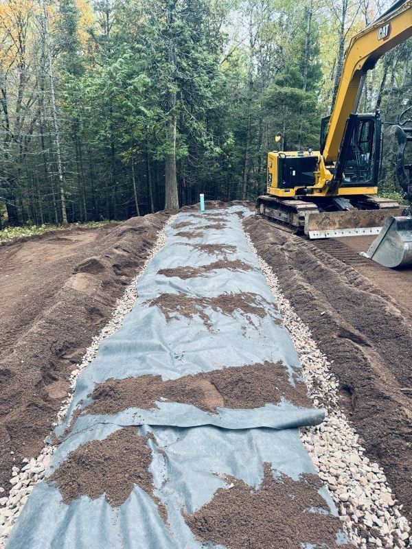 Excavator constructing a drainage system with gravel, fabric, and soil in a wooded area.