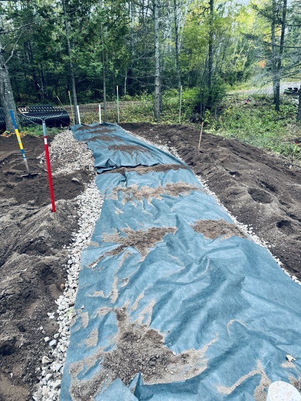 Garden bed construction: black weed barrier with soil patches, lined with gravel, in a wooded area.