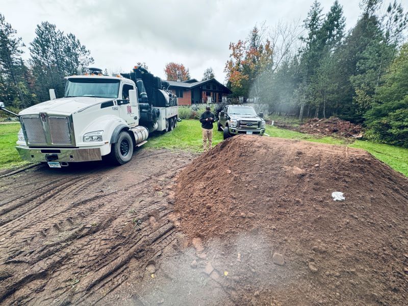 Muddy yard with a white truck, a wood chipper, a man, and a pile of dirt. A house and trees are in the background.